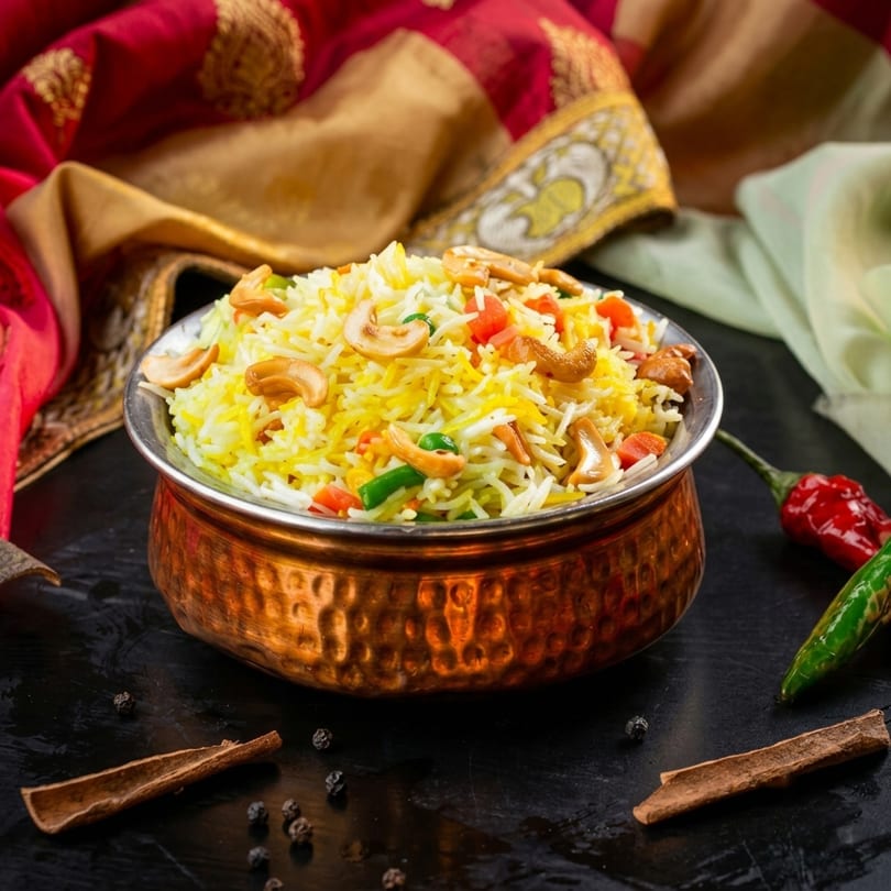Colorful rice pilaf with cashews and vegetables in a copper bowl, with spices and fabric.