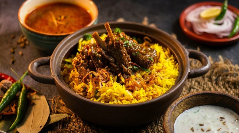 Lamb biryani in a clay pot, with side dishes of curry, raita, and sliced onions.