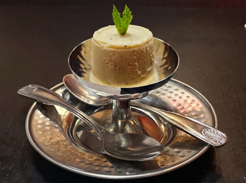 A cylindrical dessert with a mint leaf on top, served in a silver bowl with two spoons.