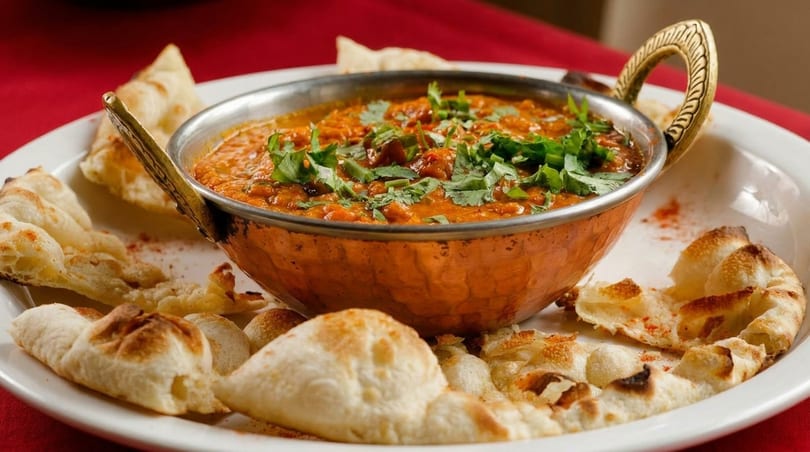 A copper bowl of Indian curry with cilantro, surrounded by pieces of naan bread on a white plate.
