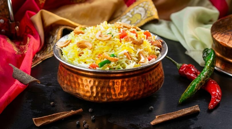 A copper bowl filled with yellow rice, cashews, and vegetables, with spices and chilies nearby.