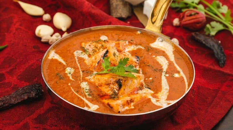 Paneer butter masala in a brass pan, garnished with cream and cilantro on a red cloth.