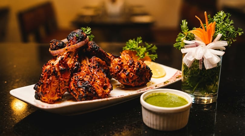 Grilled chicken drumsticks with red onion, lemon, parsley, and green chutney on a dark table.