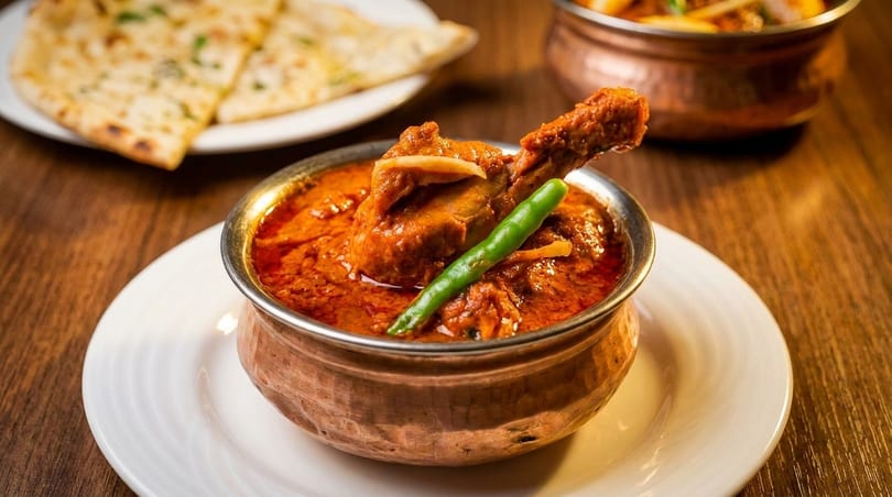 Chicken curry with a green chili in a copper bowl, with naan bread in the background.